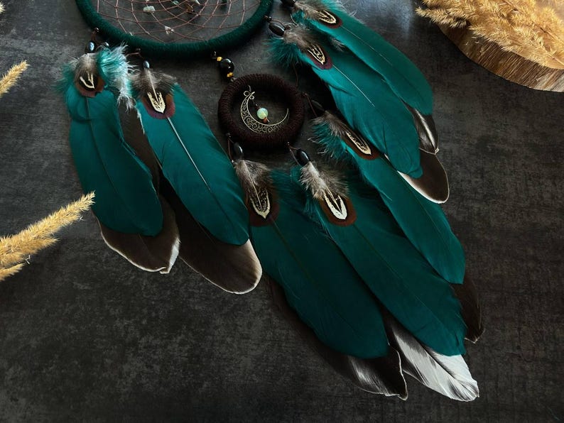 May include: A teal dreamcatcher with large, vibrant teal feathers, accented with brown and white tips. A circular pendant with a crescent moon and a small, light-colored stone hangs in the center. The dreamcatcher is a decorative item.