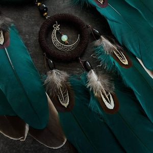 May include: A close-up of a handcrafted dreamcatcher with teal feathers and a crescent moon charm. The dreamcatcher features a woven brown ring, black beads, and detailed feather accents. The feathers have brown and white markings.