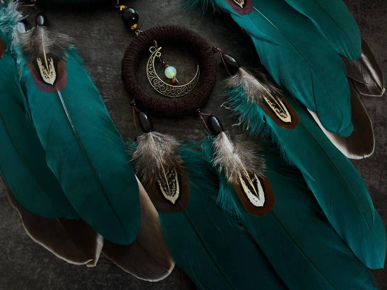 May include: A close-up of a handcrafted dreamcatcher with teal feathers and a crescent moon charm. The dreamcatcher features a woven brown ring, black beads, and detailed feather accents. The feathers have brown and white markings.