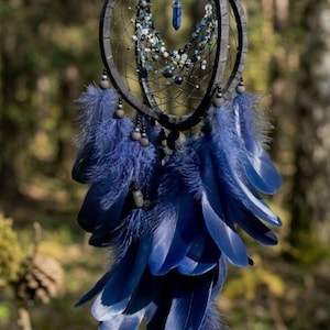 Puede incluir: Un atrapasue&ntilde;os artesanal con un dise&ntilde;o de aro negro, adornado con plumas azules, cuentas y un colgante de cristal. El atrapasue&ntilde;os est&aacute; suspendido de una rama de &aacute;rbol, mostrando sus intrincados detalles y est&eacute;tica natural.