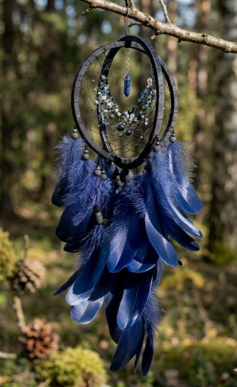 Puede incluir: Un atrapasue&ntilde;os artesanal con un dise&ntilde;o de aro negro, adornado con plumas azules, cuentas y un colgante de cristal. El atrapasue&ntilde;os est&aacute; suspendido de una rama de &aacute;rbol, mostrando sus intrincados detalles y est&eacute;tica natural.