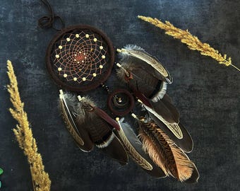 Puede incluir: Un atrapasueños marrón que presenta un aro circular grande con una red tejida, pequeñas cuentas claras y un segundo aro más pequeño. El diseño está adornado con varias plumas marrones, beige y grises, junto con un pequeño dije de media luna.