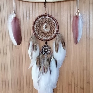 May include: A brown dream catcher with white feathers hanging from a wooden branch. The dream catcher has a crescent moon charm and a small beaded circle in the center.