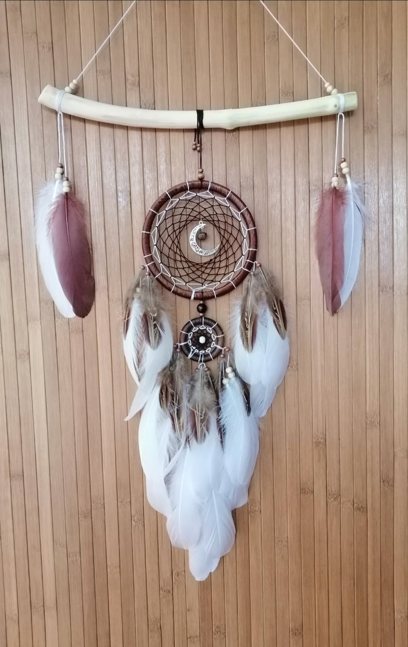 May include: A brown dream catcher with white feathers hanging from a wooden branch. The dream catcher has a crescent moon charm and a small beaded circle in the center.