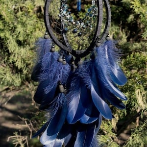 Puede incluir: Un atrapasue&ntilde;os azul oscuro con un marco circular y un intrincado dise&ntilde;o de telara&ntilde;a. Cuenta con plumas colgantes de color azul oscuro, cuentas y un colgante de cristal azul. El atrapasue&ntilde;os est&aacute; suspendido sobre un fondo de follaje verde.