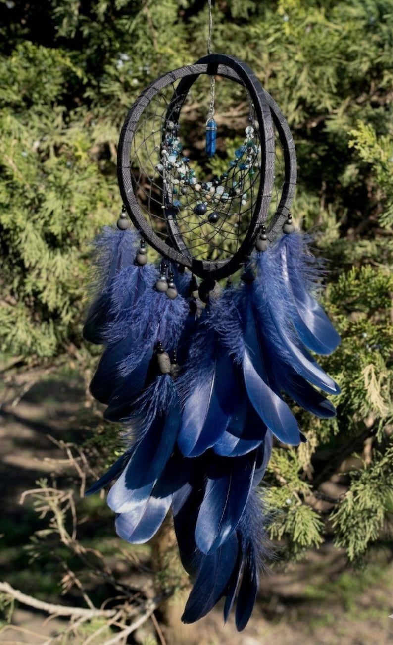 Puede incluir: Un atrapasue&ntilde;os azul oscuro con un marco circular y un intrincado dise&ntilde;o de telara&ntilde;a. Cuenta con plumas colgantes de color azul oscuro, cuentas y un colgante de cristal azul. El atrapasue&ntilde;os est&aacute; suspendido sobre un fondo de follaje verde.