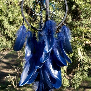 Puede incluir: Un atrapasue&ntilde;os artesanal con un marco circular gris oscuro y una red de hilo negro. Cuenta con plumas azules vibrantes y cuentas decorativas en tonos azules y verdes, colgando sobre un fondo de follaje verde.