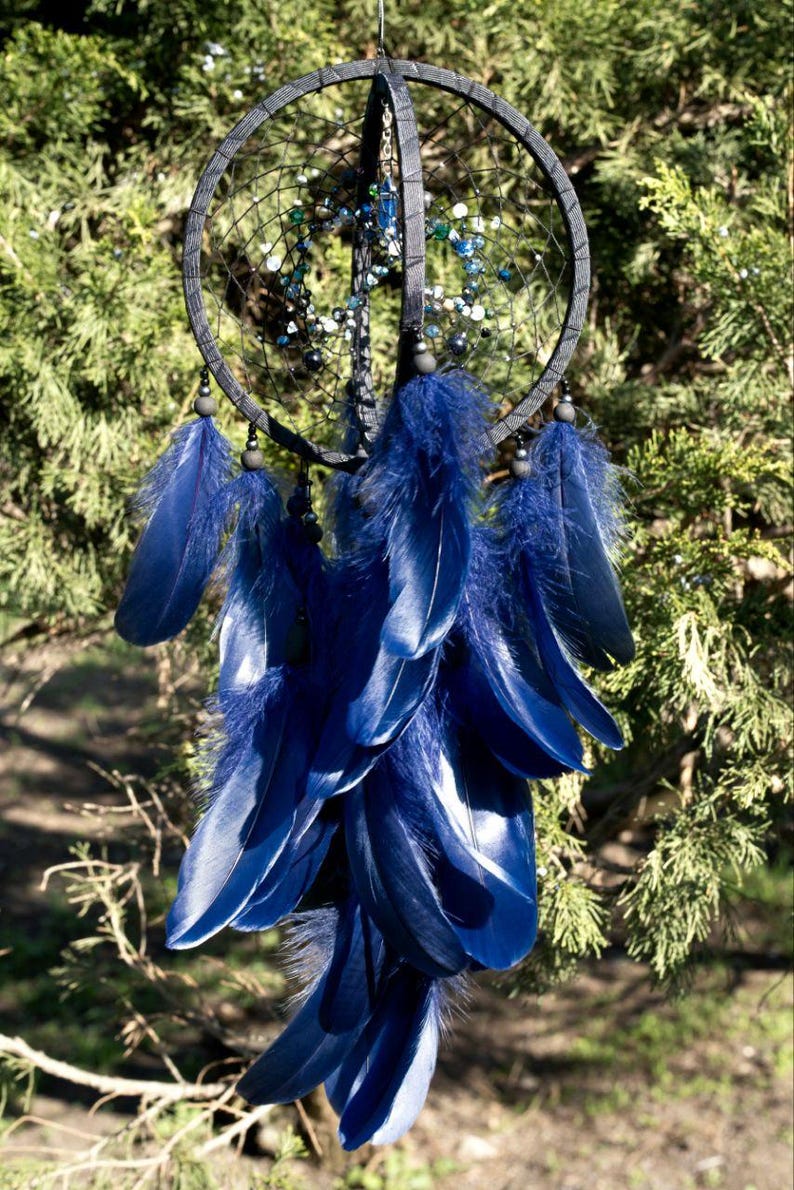Puede incluir: Un atrapasue&ntilde;os artesanal con un marco circular gris oscuro y una red de hilo negro. Cuenta con plumas azules vibrantes y cuentas decorativas en tonos azules y verdes, colgando sobre un fondo de follaje verde.