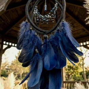 Puede incluir: Un atrapasue&ntilde;os artesanal con un marco en forma de coraz&oacute;n gris oscuro, adornado con plumas y cuentas azules. El atrapasue&ntilde;os cuelga de una estructura de madera, con un fondo natural al aire libre. Las plumas son de un azul profundo.
