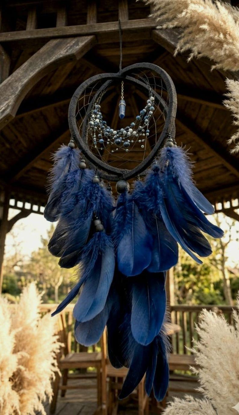 Puede incluir: Un atrapasue&ntilde;os artesanal con un marco en forma de coraz&oacute;n gris oscuro, adornado con plumas y cuentas azules. El atrapasue&ntilde;os cuelga de una estructura de madera, con un fondo natural al aire libre. Las plumas son de un azul profundo.