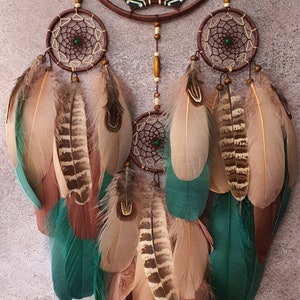 May include: A brown dreamcatcher with green and white accents and feathers. The dreamcatcher has three smaller circles with beads and feathers hanging from them.