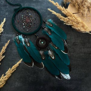 May include: A handcrafted dreamcatcher with a dark teal hoop and intricate web design. Embellished with small stones and teal feathers with white and black tips. A dark brown circular accent with a crescent moon charm is also included.