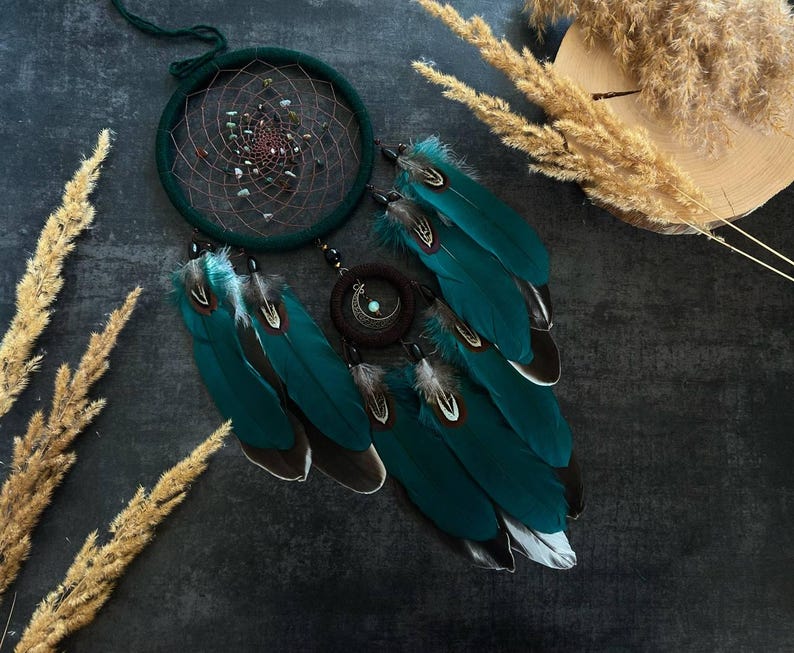 May include: A handcrafted dreamcatcher with a dark teal hoop and intricate web design. Embellished with small stones and teal feathers with white and black tips. A dark brown circular accent with a crescent moon charm is also included.