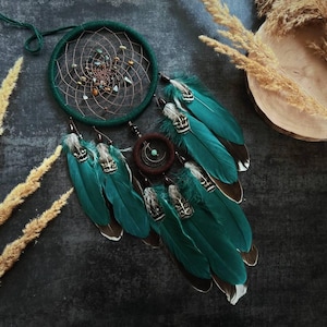 Può includere: Un acchiappasogni fatto a mano con un cerchio verde scuro e un intricato design a ragnatela. Piume verde acqua con accenti decorativi pendono sotto. L'acchiappasogni è adornato con piccole perle ed elementi naturali.