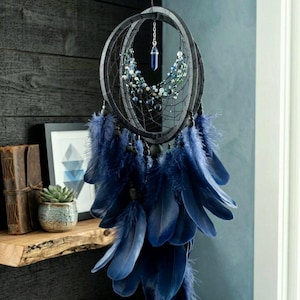Puede incluir: Un atrapasue&ntilde;os artesanal con un marco negro, adornado con plumas y cuentas azules. Un cristal azul cuelga del centro. El atrapasue&ntilde;os est&aacute; suspendido frente a una pared azul oscuro.