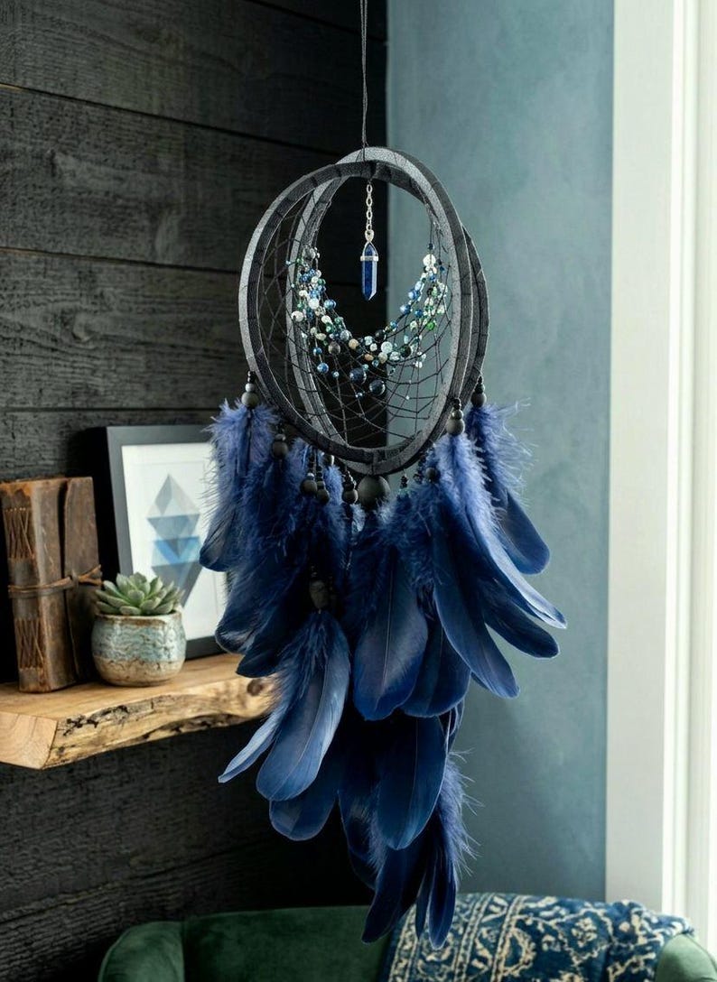 Puede incluir: Un atrapasue&ntilde;os artesanal con un marco negro, adornado con plumas y cuentas azules. Un cristal azul cuelga del centro. El atrapasue&ntilde;os est&aacute; suspendido frente a una pared azul oscuro.