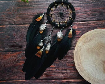 Puede incluir: Atrapasueños negro con detalles dorados y plumas negras. El atrapasueños tiene un borde de cuentas negras y una red negra y dorada. Las plumas son largas y fluidas.