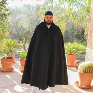 May include: A man wearing a black hooded cloak, standing in front of a potted cactus and other plants. He is wearing white socks and white shoes.