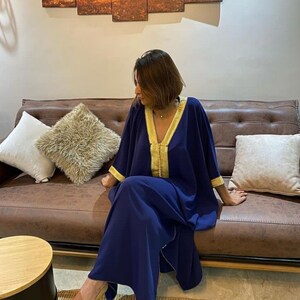 May include: A navy blue kaftan with gold trim. The kaftan has a V-neck and long sleeves. The model is sitting on a brown leather couch.