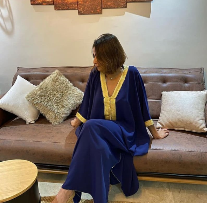May include: A navy blue kaftan with gold trim. The kaftan has a V-neck and long sleeves. The model is sitting on a brown leather couch.