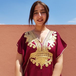 Op de afbeelding: Een vrouw draagt een bordeauxrode kaftan met gouden borduurwerk. De kaftan heeft een V-hals en korte mouwen.