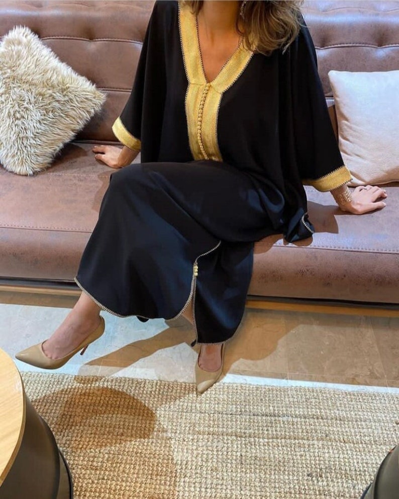 May include: A black kaftan with gold trim and a zipper down the front. The kaftan is worn by a person sitting on a brown leather couch.