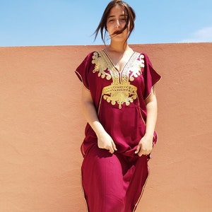 Op de afbeelding: Een maronkleurige kaftan met gouden borduurwerk. De kaftan heeft een V-hals en korte mouwen. Het borduurwerk is ingewikkeld en heeft bloem- en geometrische patronen.