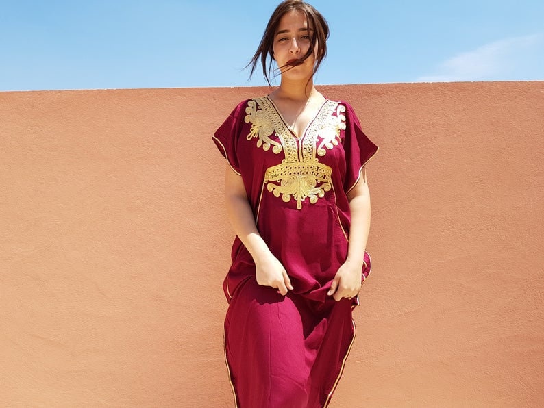 Op de afbeelding: Een maronkleurige kaftan met gouden borduurwerk. De kaftan heeft een V-hals en korte mouwen. Het borduurwerk is ingewikkeld en heeft bloem- en geometrische patronen.