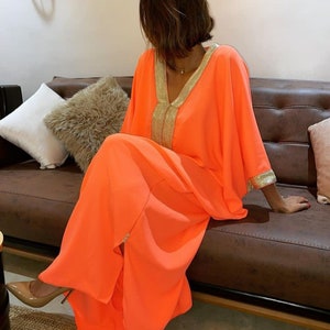 May include: A bright orange maxi dress with gold trim around the neckline and sleeves. The dress has a loose, flowing fit and a V-neckline.