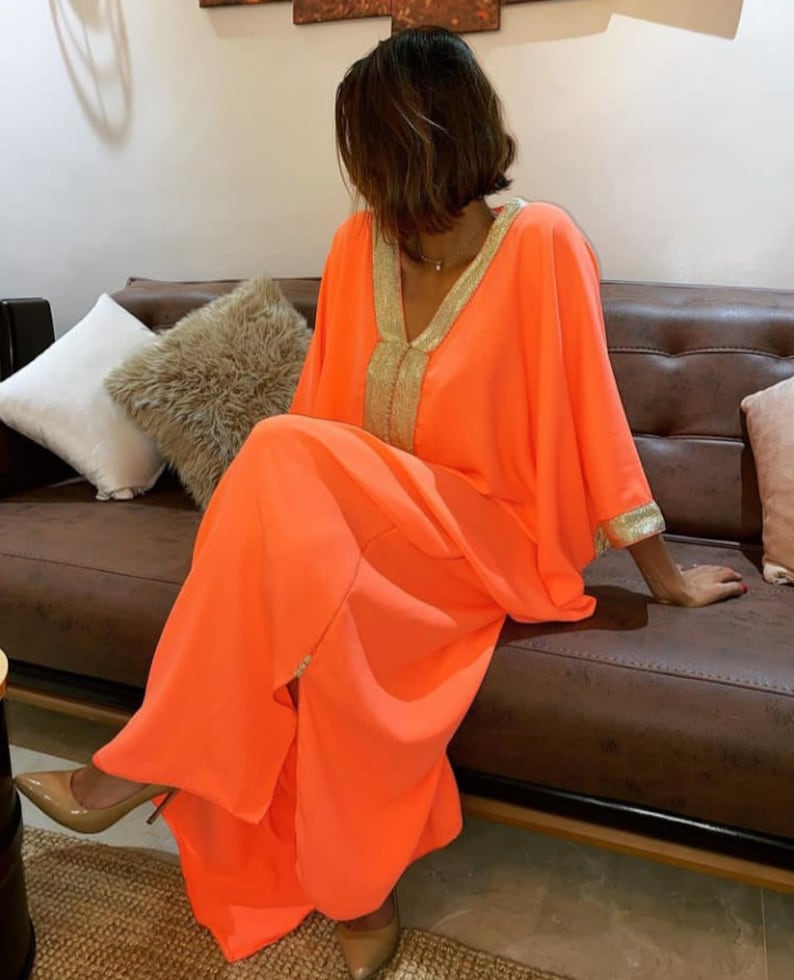 May include: A bright orange maxi dress with gold trim around the neckline and sleeves. The dress has a loose, flowing fit and a V-neckline.