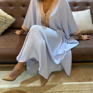 May include: A light blue kaftan with gold trim around the neckline and sleeves. The kaftan is flowing and has a relaxed fit. The model is wearing the kaftan with nude heels.