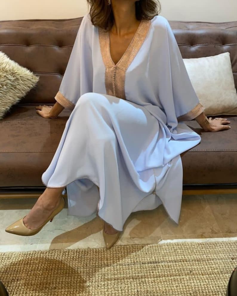 May include: A light blue kaftan with gold trim around the neckline and sleeves. The kaftan is flowing and has a relaxed fit. The model is wearing the kaftan with nude heels.