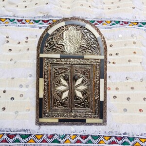 Könnte beinhalten: Ein dekorativer Bogenspiegel mit einem geschnitzten Holzrahmen und aufwendigen Metallarbeiten. Der Rahmen zeigt ein Hamsa-Hand-Design und der Spiegel ist in zwei Abschnitte unterteilt.