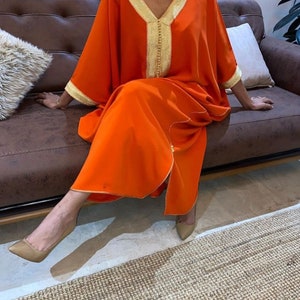 May include: A long, orange kaftan with gold trim and a V-neck. The kaftan is worn by a person sitting on a brown leather couch.