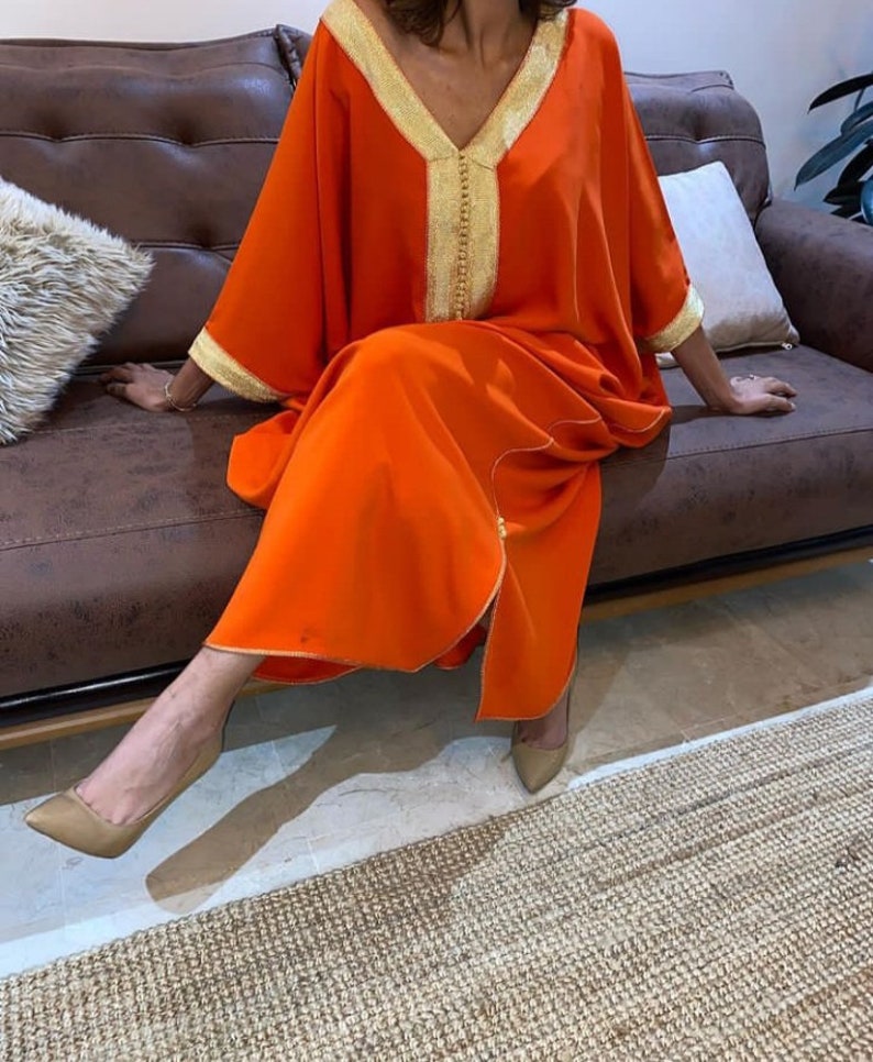 May include: A long, orange kaftan with gold trim and a V-neck. The kaftan is worn by a person sitting on a brown leather couch.
