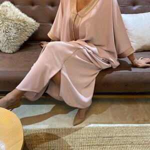 May include: A long, flowing pink kaftan with gold trim and a slit in the front. The kaftan is worn by a person sitting on a brown leather couch.