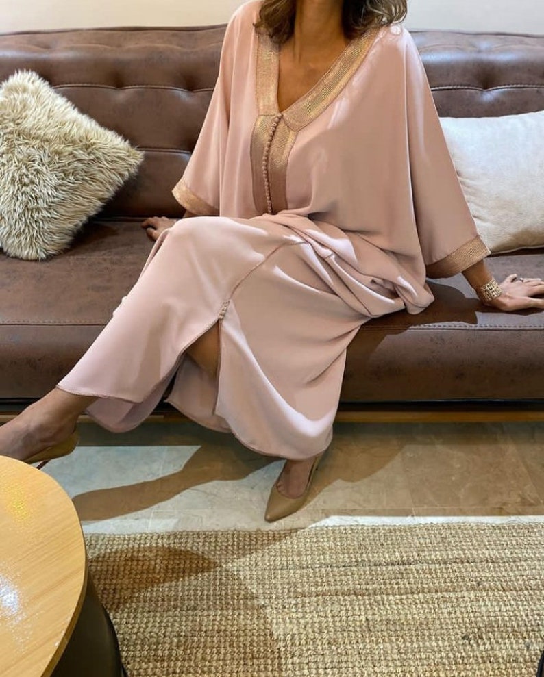 May include: A long, flowing pink kaftan with gold trim and a slit in the front. The kaftan is worn by a person sitting on a brown leather couch.
