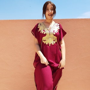Op de afbeelding: Een vrouw draagt een kastanjebruine kaftan met gouden borduurwerk. De kaftan heeft een V-hals en korte mouwen. De vrouw staat voor een perzikkleurige muur.