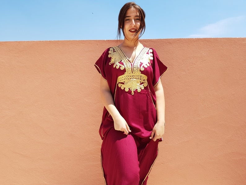 Op de afbeelding: Een vrouw draagt een kastanjebruine kaftan met gouden borduurwerk. De kaftan heeft een V-hals en korte mouwen. De vrouw staat voor een perzikkleurige muur.