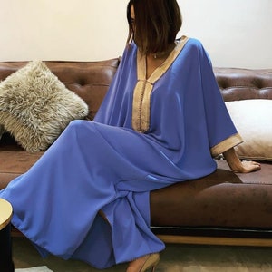 May include: A long, flowing blue kaftan with gold embroidery. The kaftan has a V-neck and long sleeves. The model is sitting on a brown leather couch.