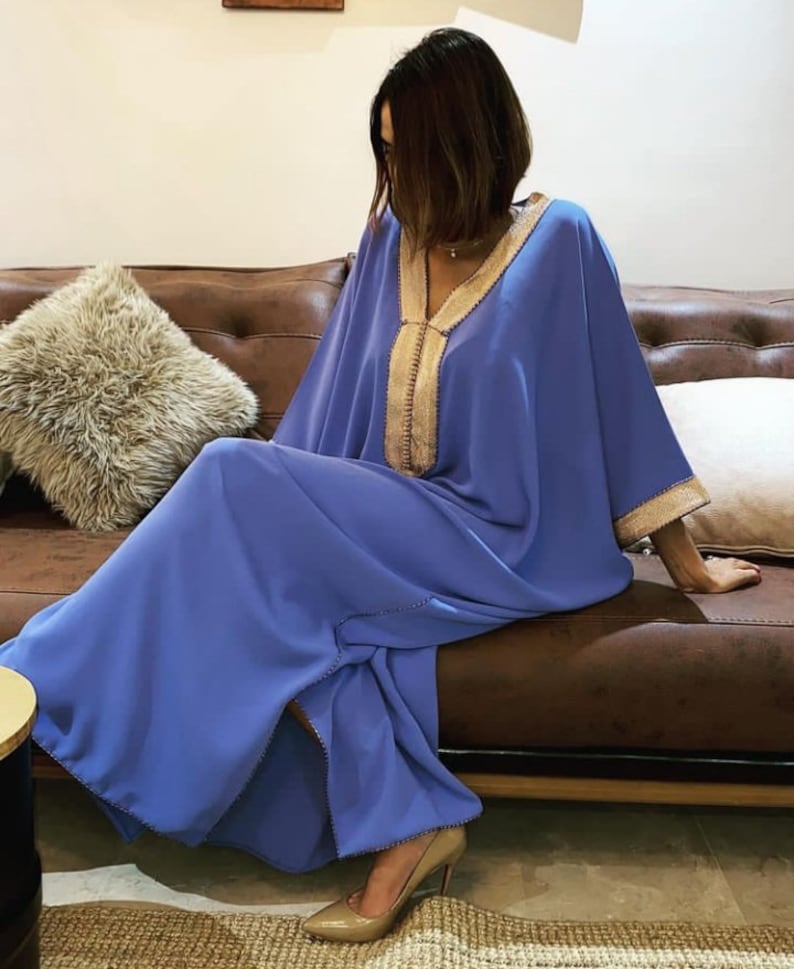 May include: A long, flowing blue kaftan with gold embroidery. The kaftan has a V-neck and long sleeves. The model is sitting on a brown leather couch.