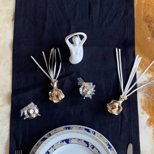 May include: A white ceramic figurine of a woman with her arms raised above her head, surrounded by three decorative floral arrangements with white ribbons and a small silver and white decorative object. A white plate with a blue and gold border sits on a black tablecloth, along with a silver fork and knife.