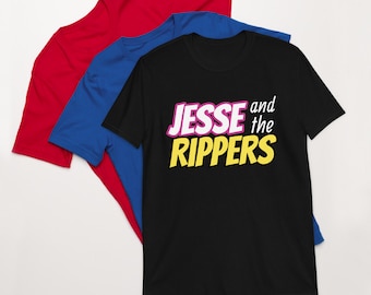 May include: Three t-shirts stacked on top of each other. The top shirt is black with the text "Jesse and the Rippers" in yellow, pink, and white letters. The middle shirt is blue and the bottom shirt is red.