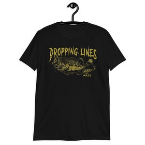 Puede incluir: Camiseta negra con un gráfico dorado de un pez de bajo y el texto "Dropping Lines Beers 'n Bass".