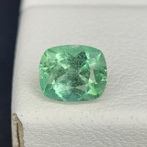 Puede incluir: Una piedra preciosa de talla cojín con un color verde claro vibrante y una superficie facetada que refleja la luz. La piedra muestra características de claridad interna y una forma simétrica con esquinas redondeadas y un aspecto pulido.