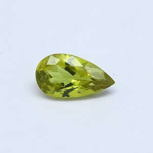 Peut inclure: Une pierre précieuse en forme de poire, facettée, d'une couleur vert-jaunâtre vibrante. La gemme est posée sur un fond blanc uni, soulignant sa clarté et sa taille. Les facettes de la pierre précieuse reflètent la lumière, créant un effet scintillant.