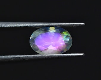 May include: An oval-cut gemstone held by tweezers against a black background. The gem displays a vibrant play of colors, including purple, green, and yellow, with a polished, iridescent surface. The gemstone is a focal point.