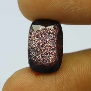 May include: A rectangular-cut gemstone with a dark brown base and a shimmering, speckled interior of pink, orange, and iridescent colours. The gem is held between two fingers, showcasing its polished facets and unique colour play.