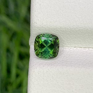 Puede incluir: Una gema verde vibrante, de talla cojín. La gema es de un color verde esmeralda intenso con facetas que reflejan la luz. La gema se encuentra sobre un fondo blanco, con un fondo verde borroso.