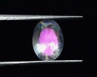 May include: A clear oval-shaped gemstone with iridescent rainbow colors and a pink center, held by tweezers against a black background.
